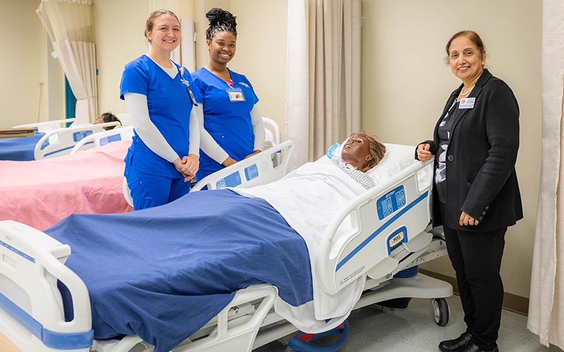 Dean Anumolu stands with two fdtc students with a medical bed and training dummy.